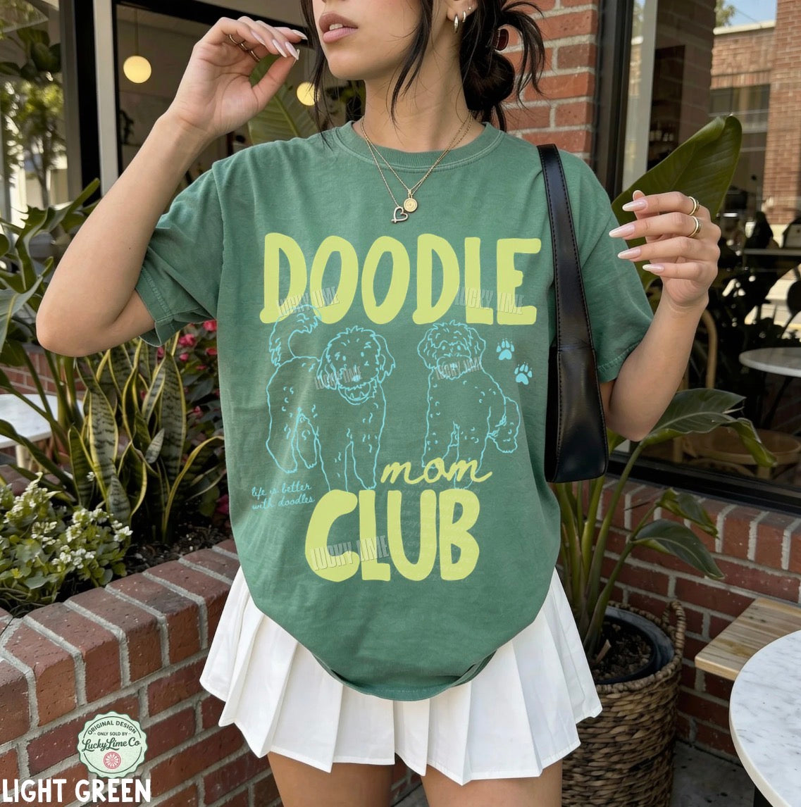 Person wearing a green t-shirt with 'Doodle Mom Club' text and graphics outdoors.