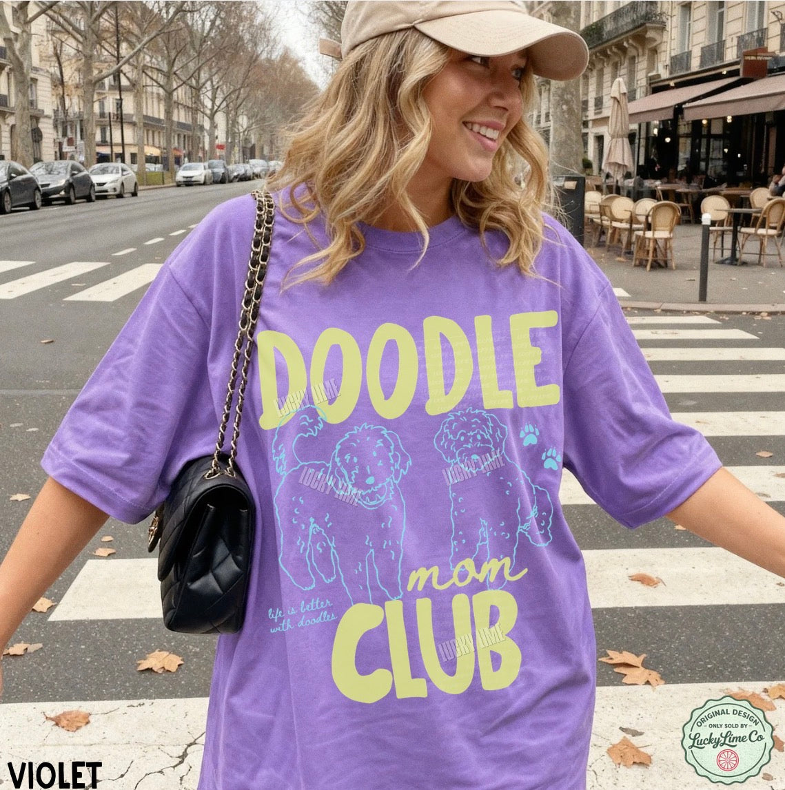 Woman wearing a purple t-shirt with 'DOODLE mom CLUB' text and graphics on a city street.