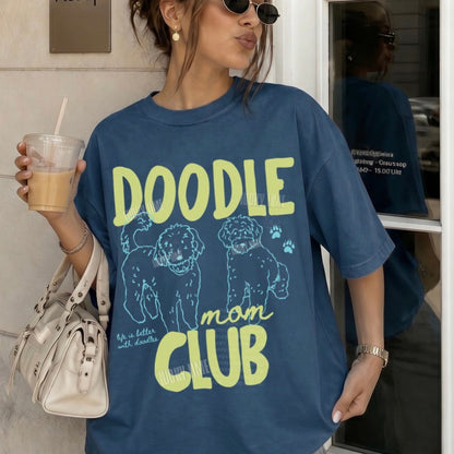 Person wearing a blue t-shirt with 'Doodle Mom Club' text and graphics, holding a drink.