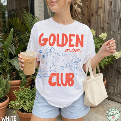 Person wearing a 'Golden Mom Club' t-shirt holding a drink outdoors.