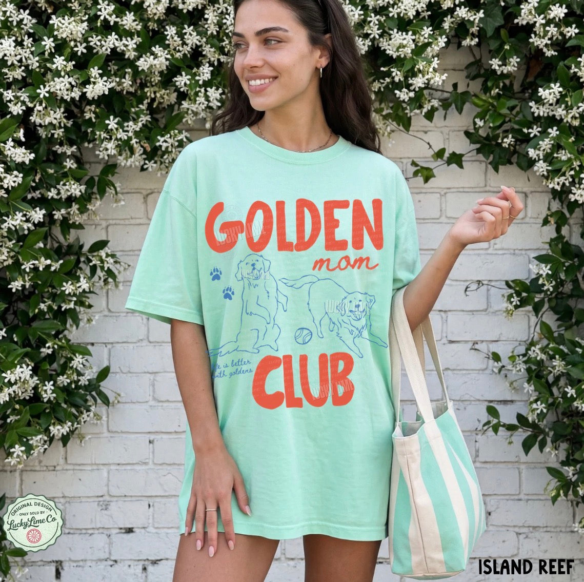 Woman wearing a light green t-shirt with 'Golden Mom Club' text and graphics, standing in front of a white brick wall with greenery.