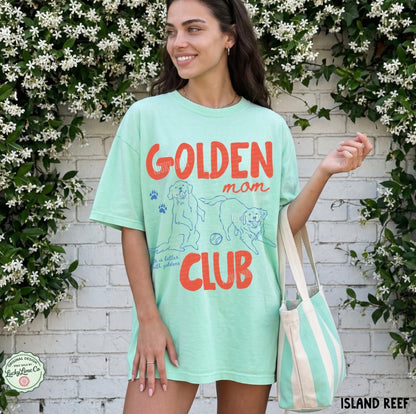 Woman wearing a light green t-shirt with 'Golden Mom Club' text and graphics, standing in front of a white brick wall with greenery.