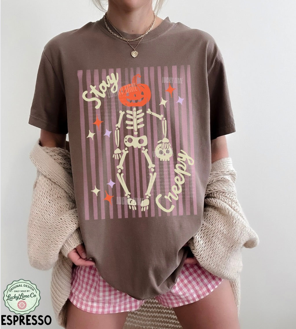Person wearing a brown t-shirt with a skeleton design and 'Stay Creepy' text, holding a beige sweater.