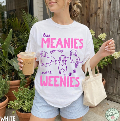 Person wearing a white t-shirt with pink and purple text and graphics, holding a drink and a bag outdoors.