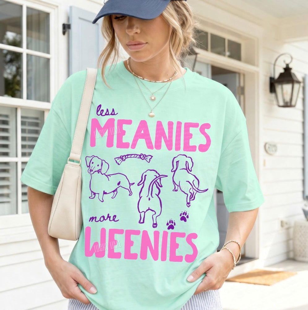 Woman wearing a light green t-shirt with 'less MEANIES more WEENIES' text and dog illustrations, standing outdoors.