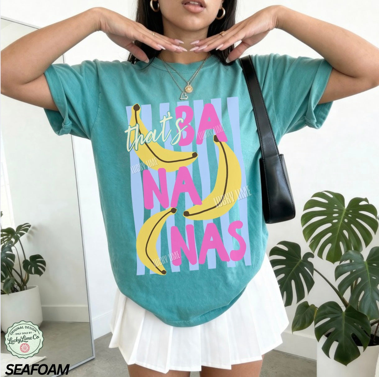Person wearing a teal t-shirt with banana graphic and text, standing indoors with plants in the background.