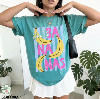 Person wearing a teal t-shirt with banana graphic and text, standing indoors with plants in the background.