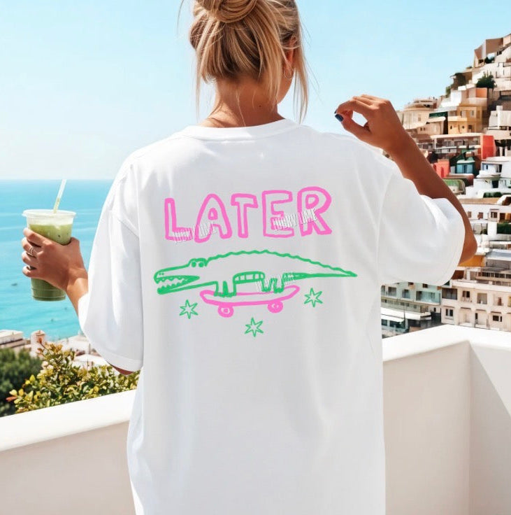 Person wearing a white t-shirt with 'LATER' and alligator design, holding a drink, with a scenic background.