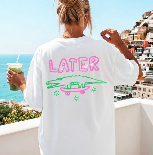 Person wearing a white t-shirt with 'LATER' and alligator design, holding a drink, with a scenic background.