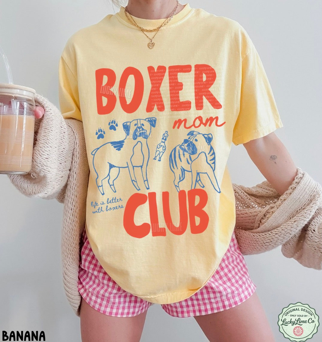 Person wearing a yellow t-shirt with 'Boxer Mom Club' design, holding a drink.