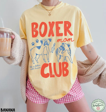 Person wearing a yellow t-shirt with 'Boxer Mom Club' design, holding a drink.