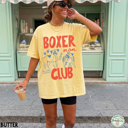 Woman wearing a yellow t-shirt with 'Boxer Man Club' text and graphics, standing in front of a green building.