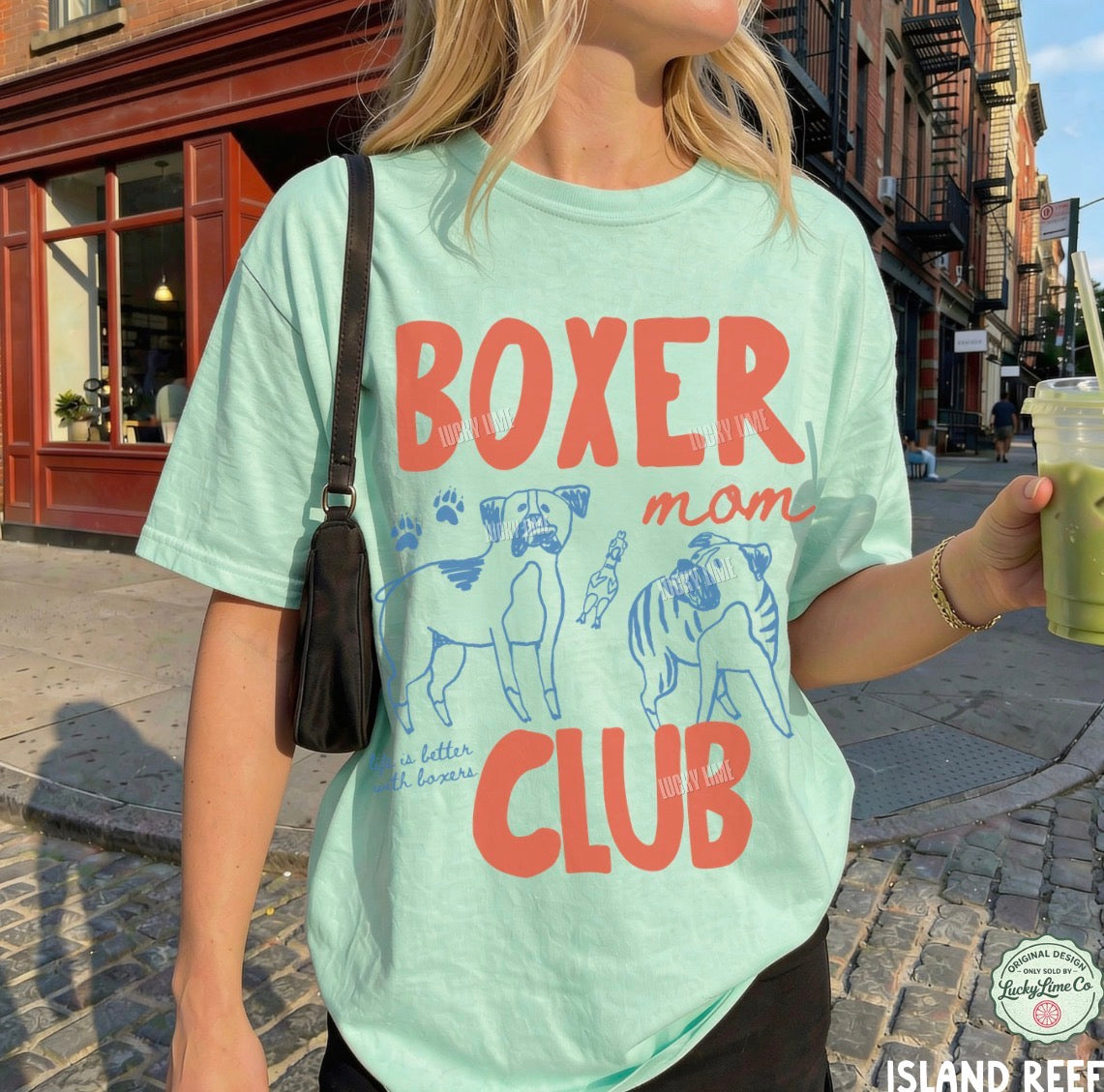 Person wearing a light green t-shirt with 'Boxer Mom Club' text and dog illustrations on a city street.