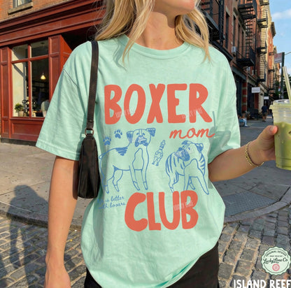 Person wearing a light green t-shirt with 'Boxer Mom Club' text and dog illustrations on a city street.