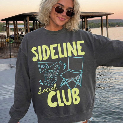 Person wearing a sweatshirt with 'Sideline Social Club' text by a lake.