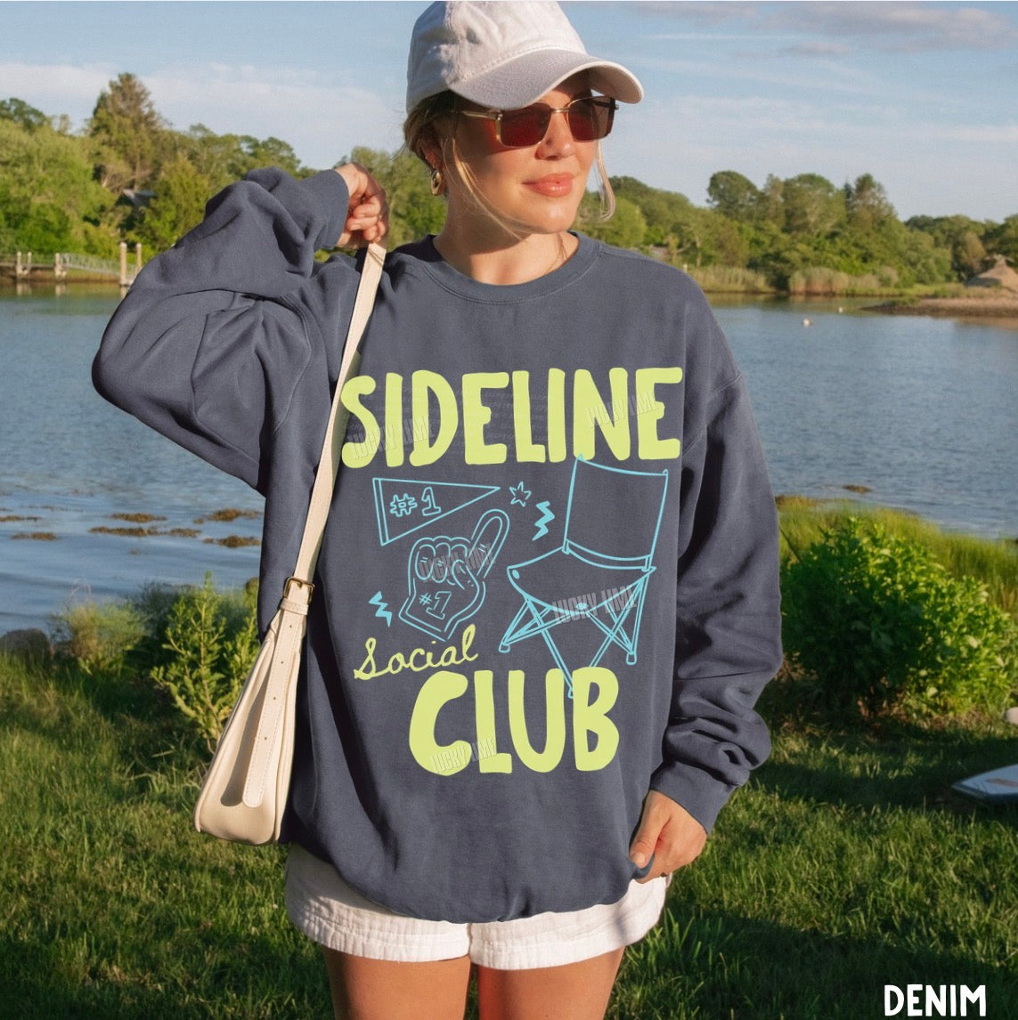 Person wearing a denim sweatshirt with 'Sideline Social Club' text by a lake.