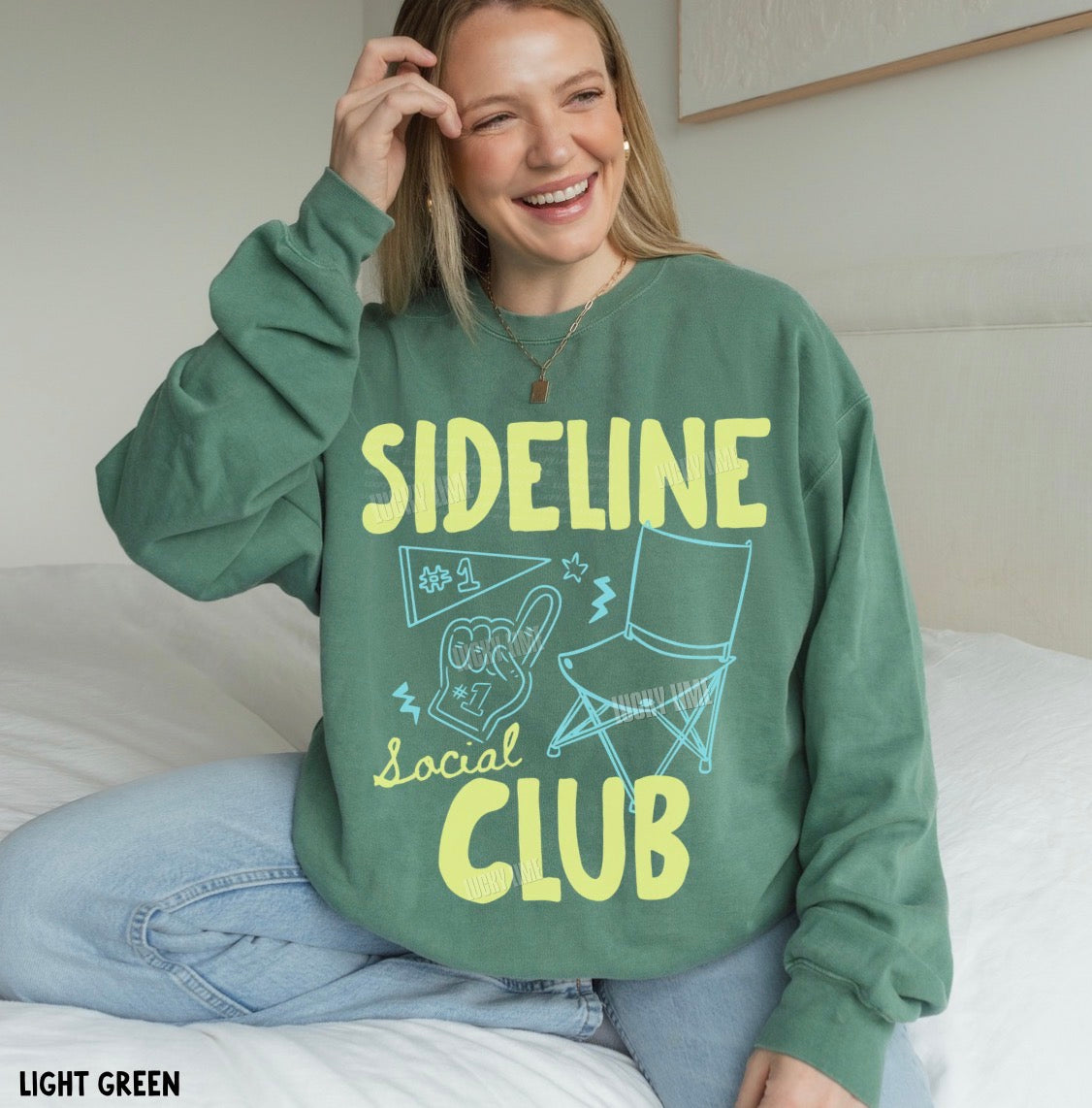 Person wearing a green sweatshirt with 'Sideline Social Club' text and graphics, sitting on a bed.