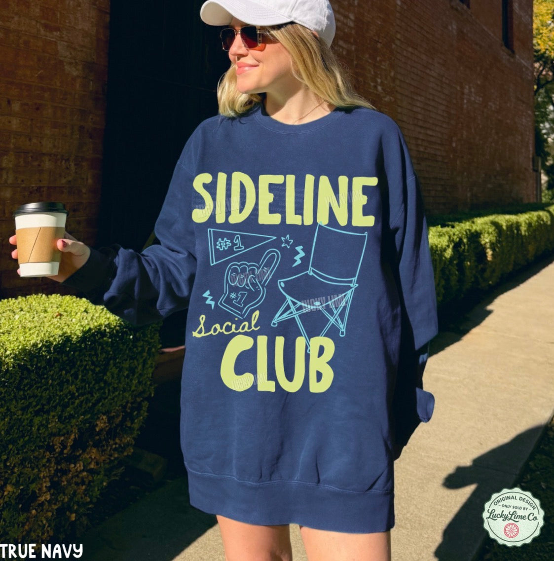 Person wearing a navy sweatshirt with 'Sideline Social Club' text and graphics, holding a coffee cup outdoors.