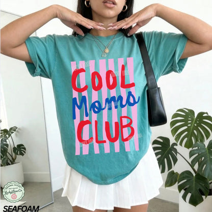 Person wearing a teal t-shirt with 'Cool Moms Club' text in a striped pattern, standing indoors with plants in the background.