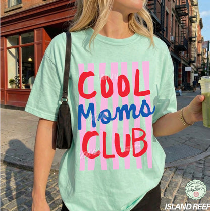 Person wearing a light green t-shirt with 'Cool Moms Club' text on a city street.