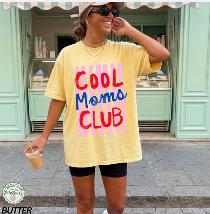 Woman wearing a yellow t-shirt with 'Cool Moms Club' text, standing in front of a light green building.