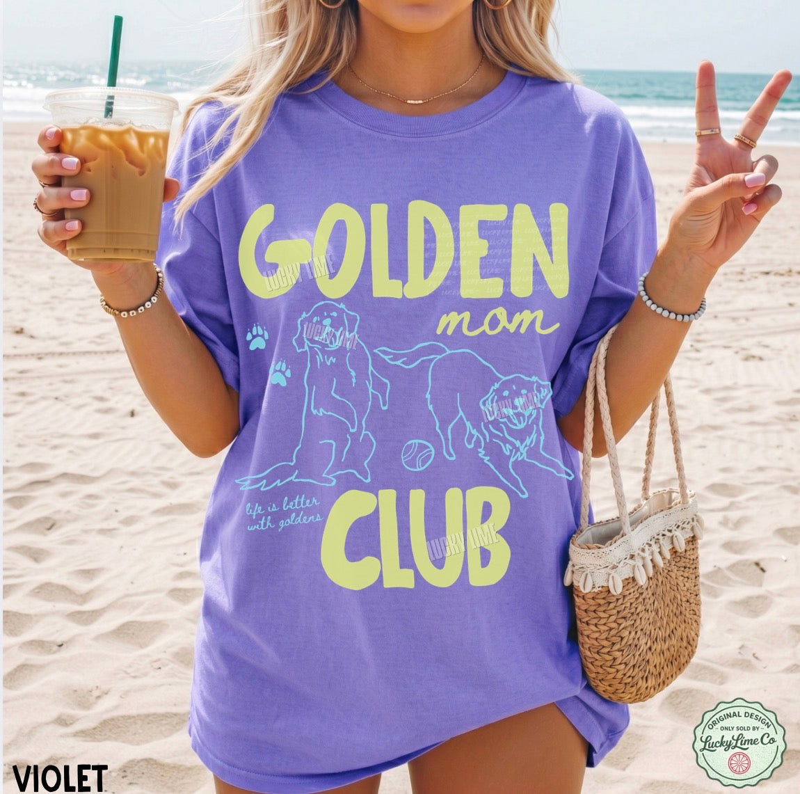 Person wearing a purple 'Golden Mom Club' t-shirt on a beach, holding a drink.