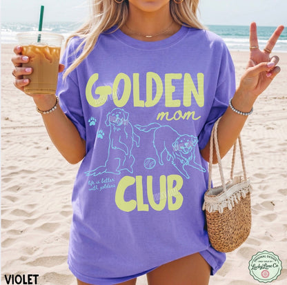 Person wearing a purple 'Golden Mom Club' t-shirt on a beach, holding a drink.