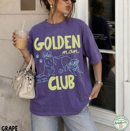 Person wearing a purple 'Golden Mom Club' t-shirt holding a drink.