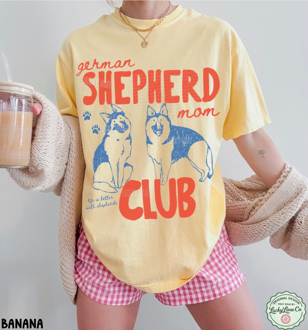 Person wearing a yellow t-shirt with 'German Shepherd Mom Club' design, holding a drink.