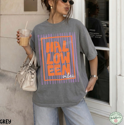 Person wearing a grey 'HALLOWEEN vibes' t-shirt holding a drink, standing in front of a building.