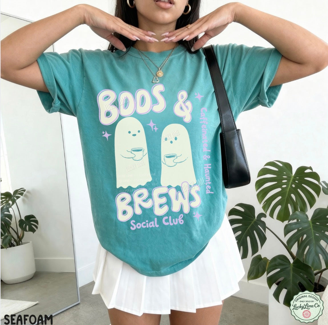 Person wearing a teal t-shirt with ghost graphics and text, standing indoors with plants in the background.
