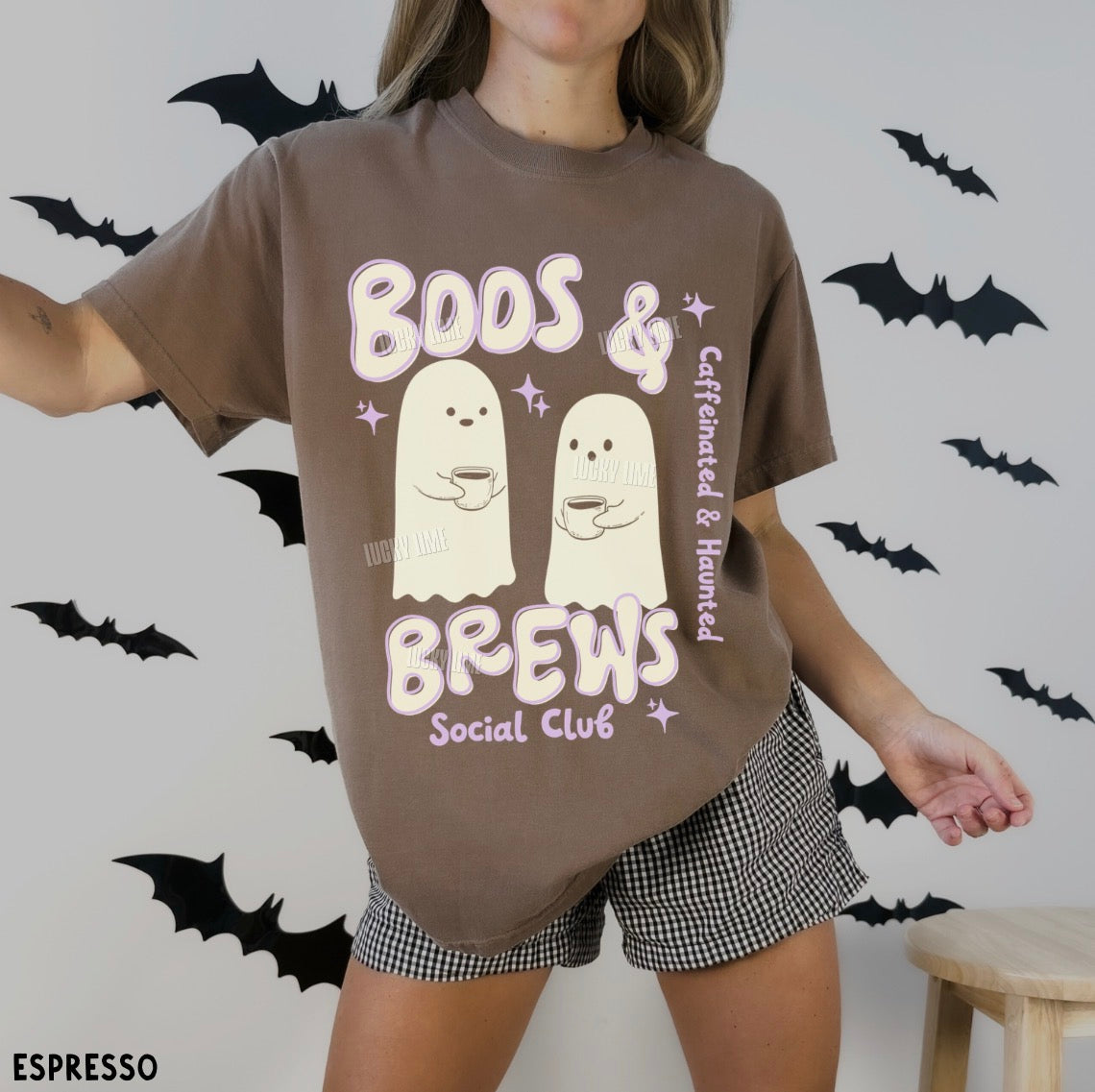 Person wearing a brown t-shirt with ghost graphics and text, standing against a wall with black bat decorations.