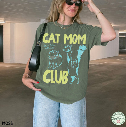 Woman wearing a green t-shirt with 'Cat Mom Club' text and graphics in an indoor setting.