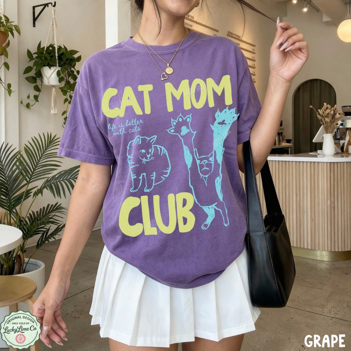 Person wearing a purple 'Cat Mom Club' t-shirt in an indoor setting