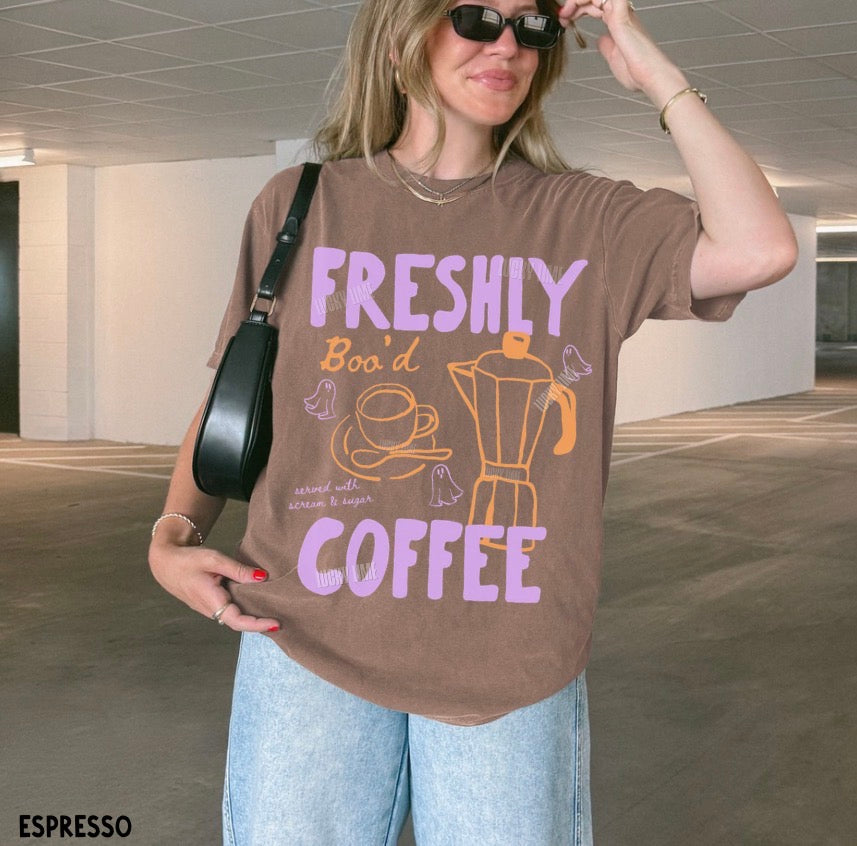 Woman wearing a brown t-shirt with 'Freshly Bod' Coffee' text and graphics in an indoor setting.