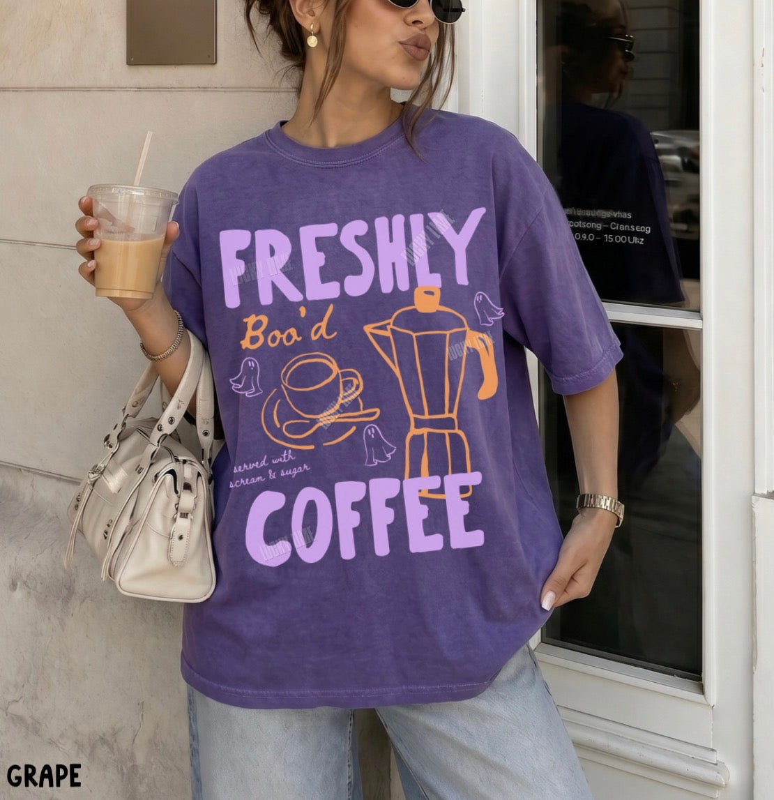 Person wearing a purple t-shirt with 'FRESHLY BOO'D COFFEE' text and graphics, holding a drink.
