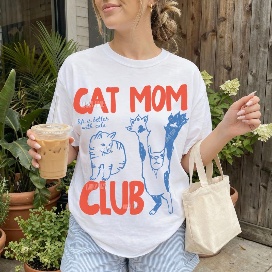 Person wearing a 'Cat Mom Club' t-shirt holding a drink outdoors.