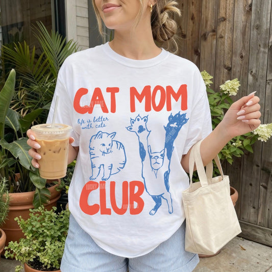 Person wearing a 'Cat Mom Club' t-shirt holding a drink outdoors.