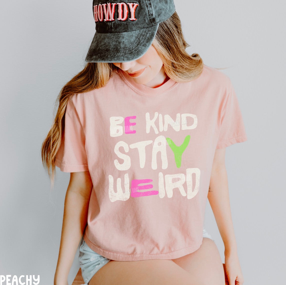 A woman wearing a light pink graphic t-shirt with the text 'BE KIND STAY WEIRD' in white, pink, and green, paired with a dark gray denim cap and denim shorts.