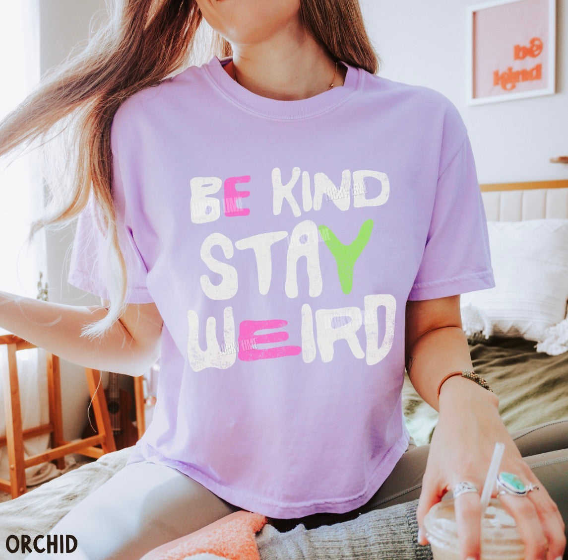 A woman wearing a lavender-purple graphic t-shirt with the text 'BE KIND STAY WEIRD' in white, pink, and green lettering. She is sitting on a bed in a bedroom setting.