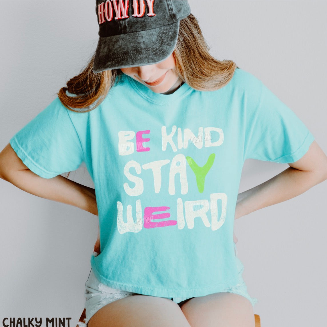 A woman wearing a light aqua graphic t-shirt with the text 'BE KIND STAY WEIRD' in white, pink, and green lettering. She is also wearing a dark gray cap with pink text and denim shorts.