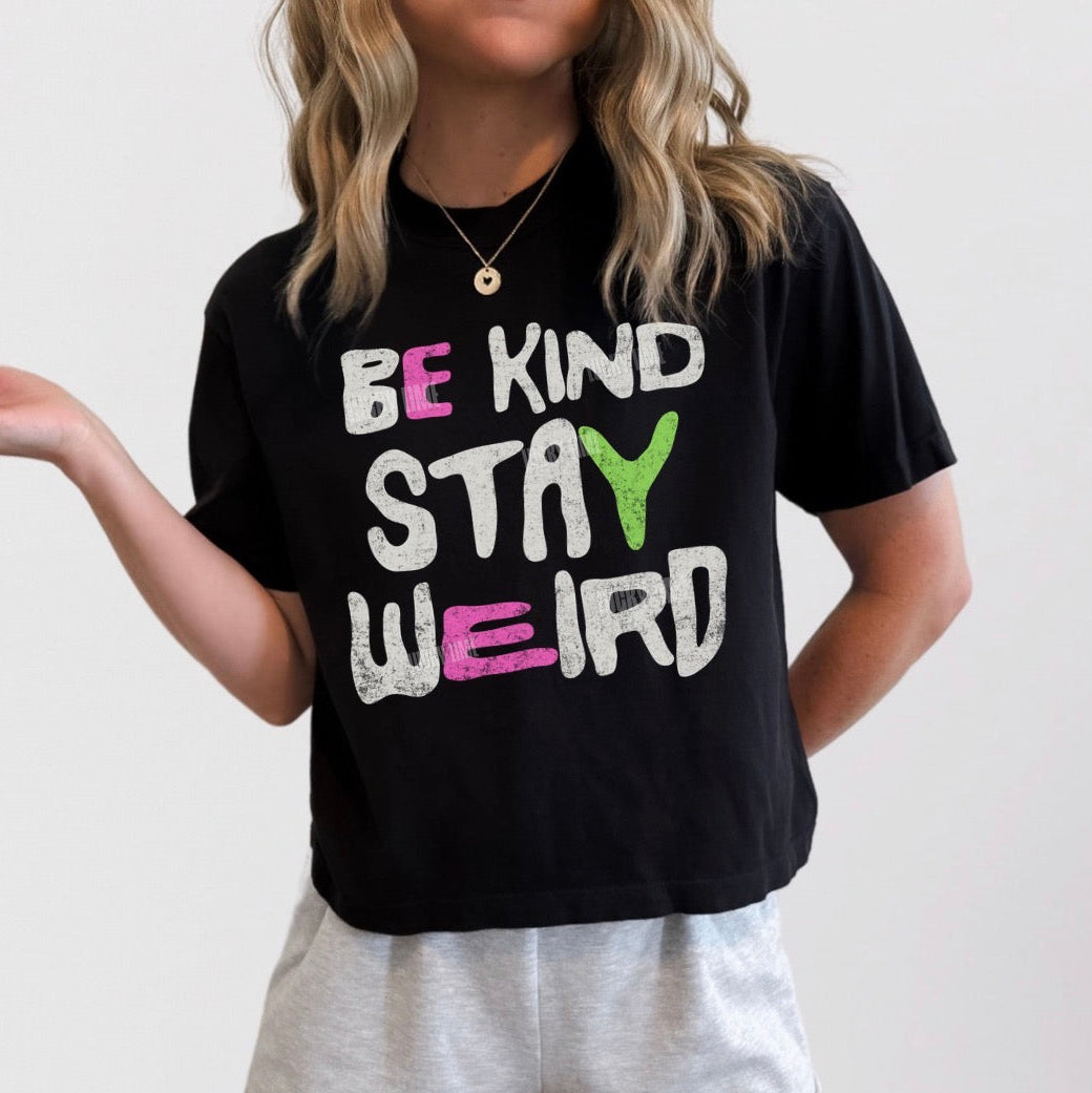 A woman wearing a black graphic t-shirt with the text 'BE KIND STAY WEIRD' in white, pink, and green, paired with light gray shorts against a plain light gray background.