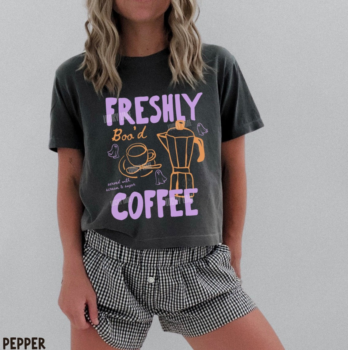 A woman wearing a dark charcoal gray graphic t-shirt with purple and yellow text reading 'FRESHLY Boo'd COFFEE' and black-and-white gingham shorts, against a plain light gray background.