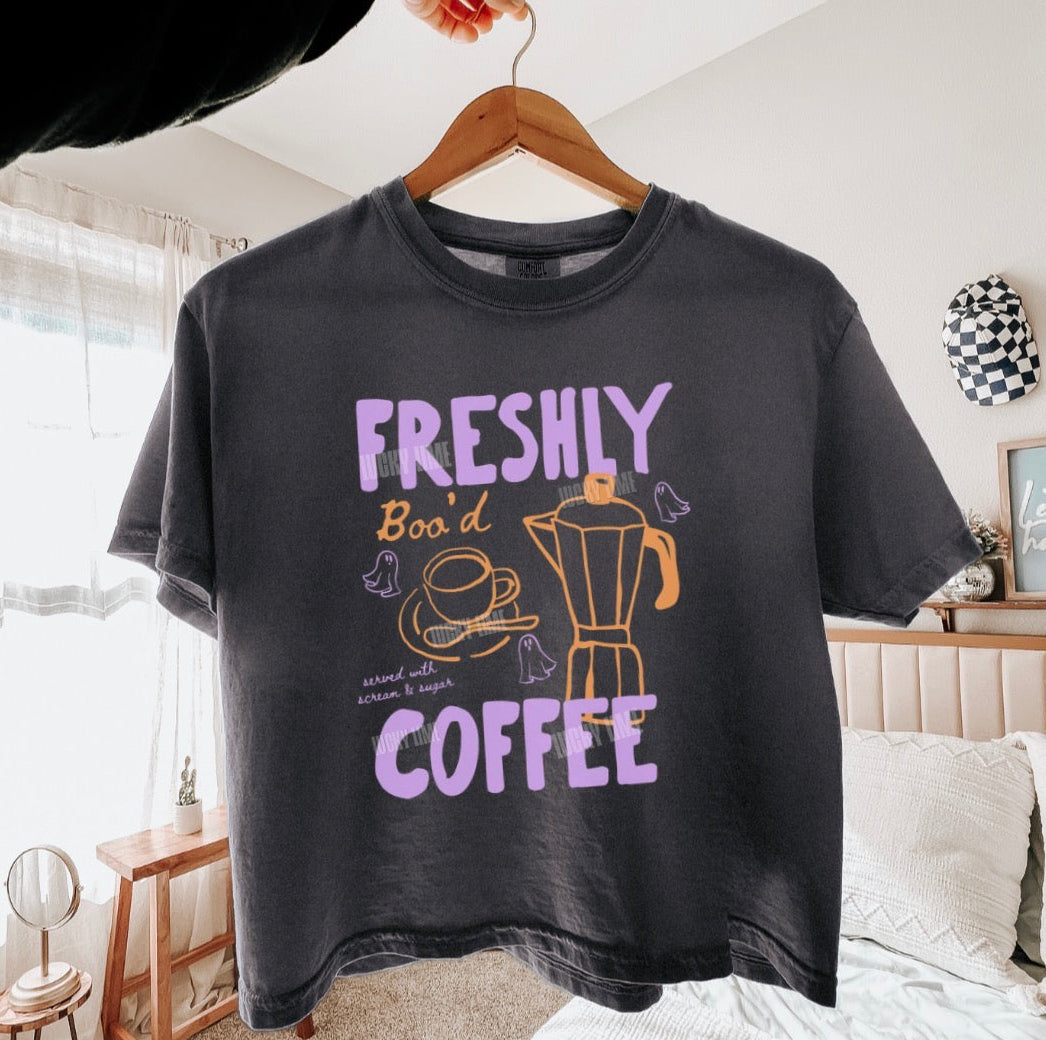 A charcoal gray graphic t-shirt hanging on a wooden hanger in a bedroom setting. The shirt features a distressed purple and yellow graphic that reads 'FRESHLY BOO'D COFFEE' with illustrations of a coffee cup, hat, and coffee maker.
