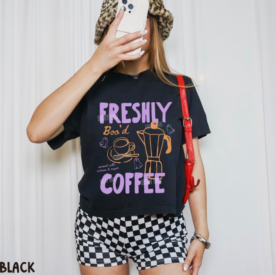 A woman takes a mirror selfie wearing a black graphic t-shirt with 'FRESHLY BOODED COFFEE' in purple and orange text, paired with black and white checkered shorts and a leopard print beanie.