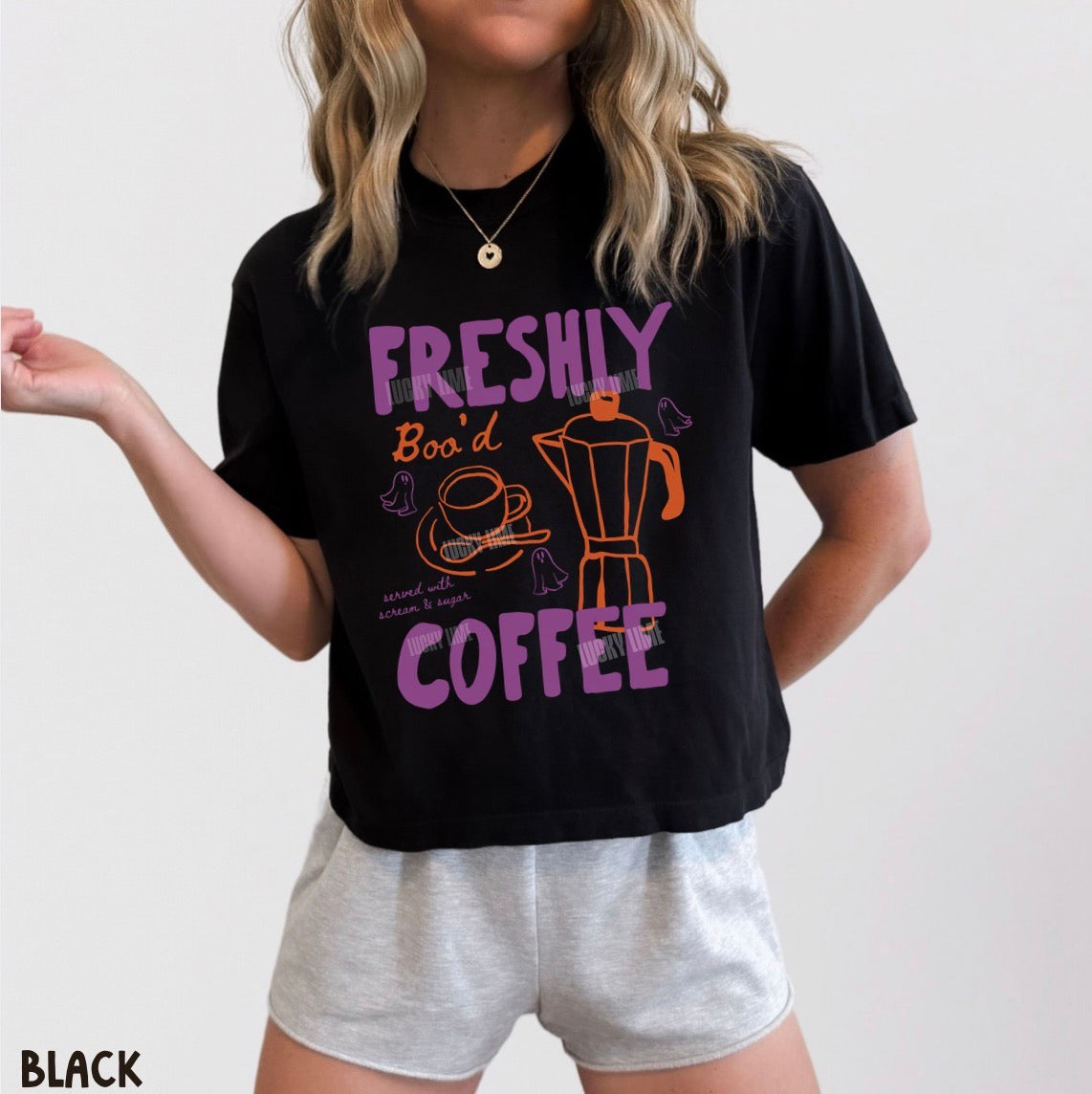 A woman wearing a black graphic t-shirt with purple and orange text reading 'FRESHLY Boo'd COFFEE' and a coffee-themed illustration, paired with light gray shorts against a plain light background.
