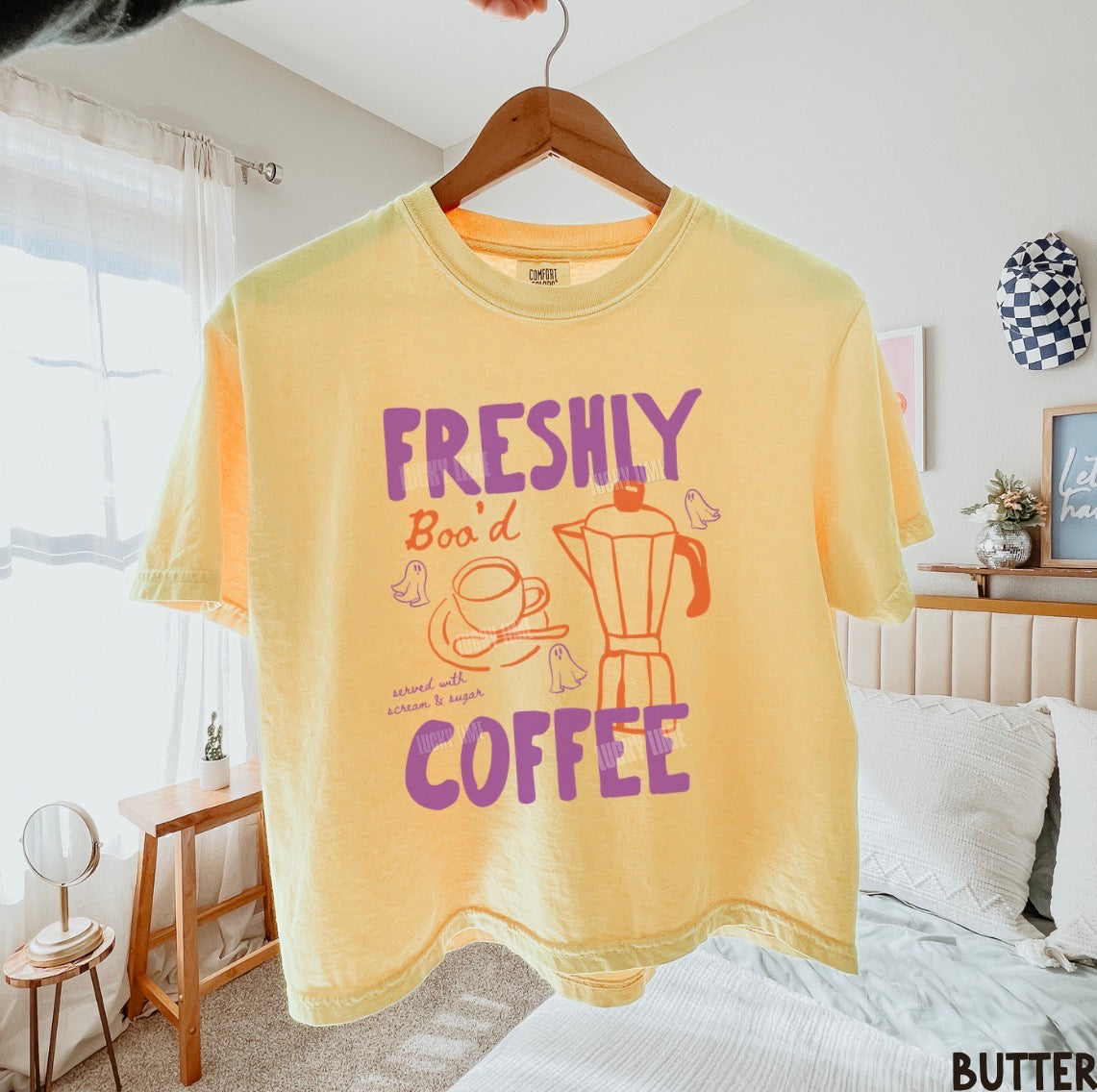 A pale yellow graphic t-shirt hanging on a wooden hanger in a bedroom setting. The shirt features a retro-style purple and orange print that reads 'FRESHLY Boo'd COFFEE' with illustrations of a coffee cup, coffee maker, and birds.
