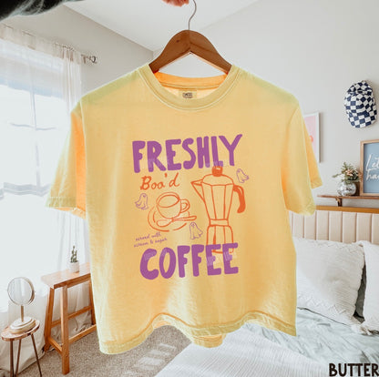 A pale yellow graphic t-shirt hanging on a wooden hanger in a bedroom setting. The shirt features a retro-style purple and orange print that reads 'FRESHLY Boo'd COFFEE' with illustrations of a coffee cup, coffee maker, and birds.
