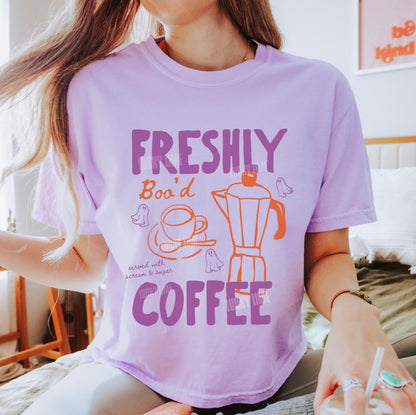 A woman wearing a light purple graphic t-shirt with the words 'FRESHLY BOO D COFFEE' and illustrations of a coffee cup and coffee maker. The photo is taken in a brightly lit bedroom setting, with the brand name 'ORCHID' visible in the bottom-left corner.