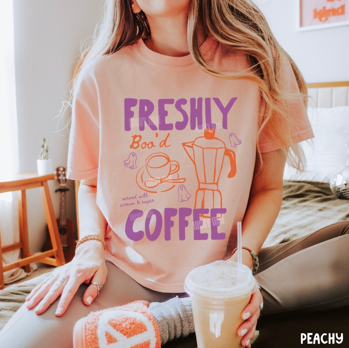 A woman wearing a peach-pink graphic t-shirt with purple and orange text reading 'FRESHLY Bo'oid COFFEE' and an illustration of a coffee mug and moka pot, sitting on a bed in a cozy bedroom setting.
